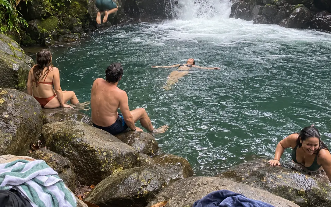 Friends at a natural pool below a waterfall in the rainforest, one jumping in, one floating
