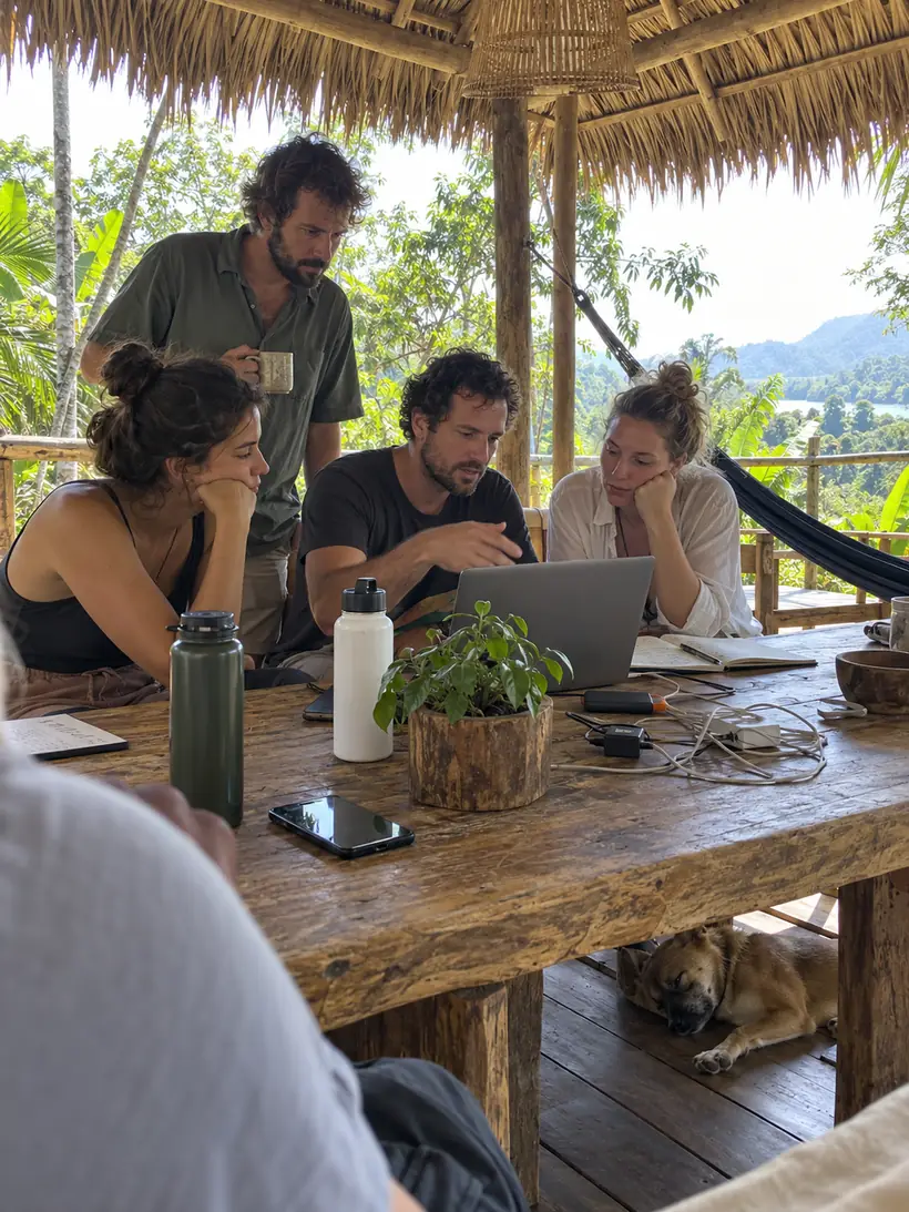 Four creators around one laptop at a wooden table under a thatched roof, one explaining, a dog asleep underneath