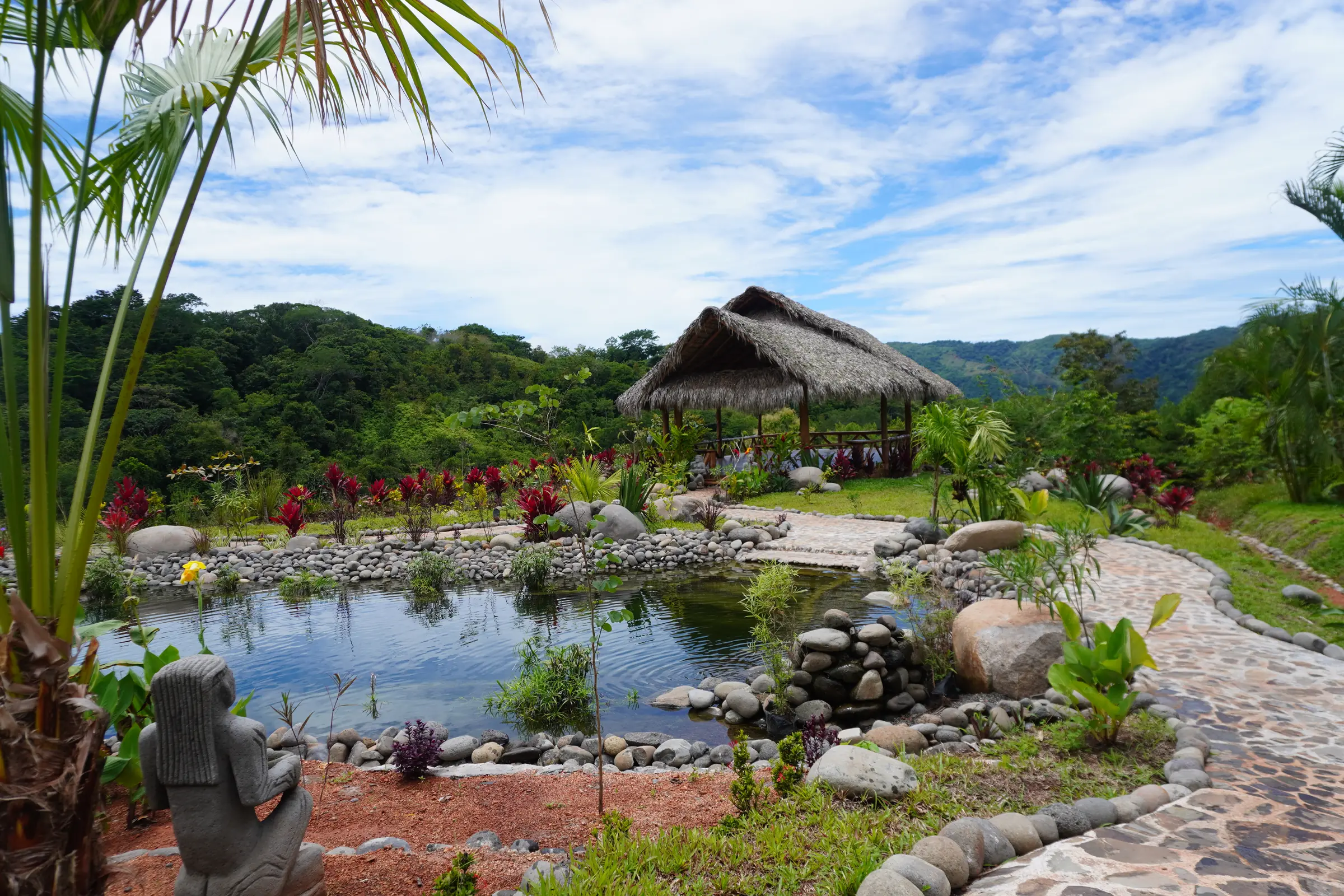 A natural pool among flowers and stones, with the yoga shala and mountains behind